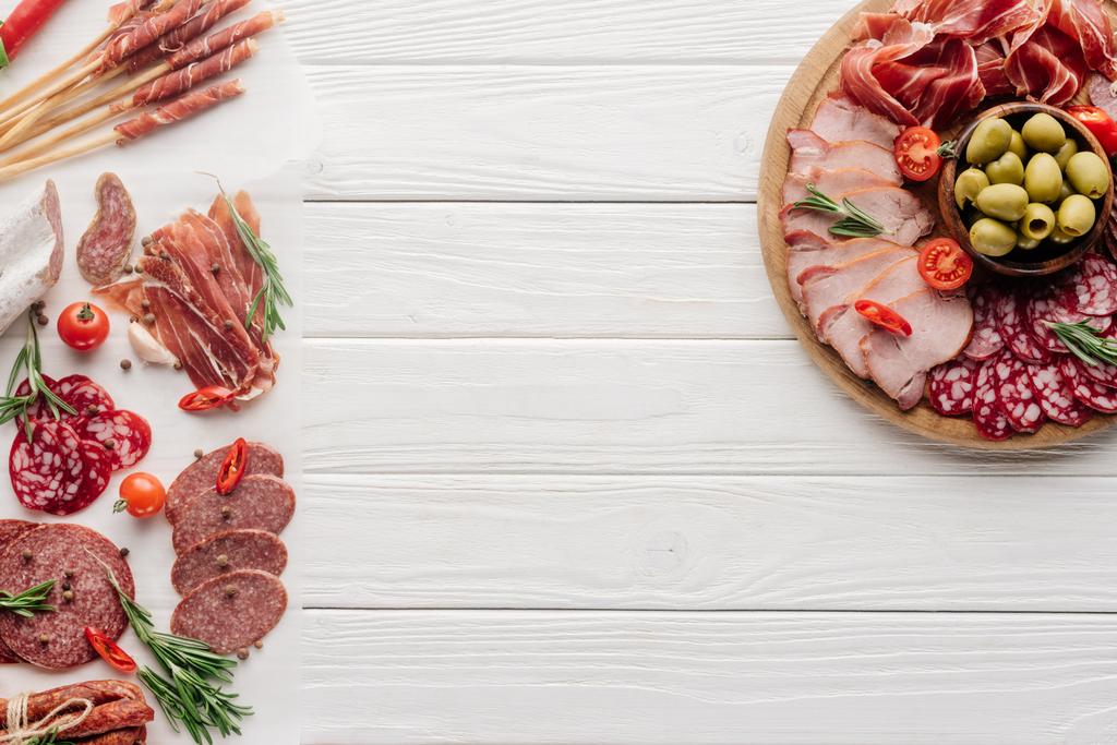 Draufsicht auf die Anordnung verschiedener Fleischsnacks und Oliven auf weißem Holzhintergrund - Foto, Bild