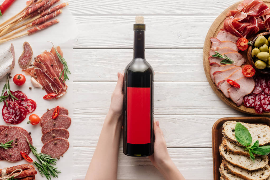 partial view of woman holding bottle of red wine on white tabletop with meat appetizers - Photo, Image