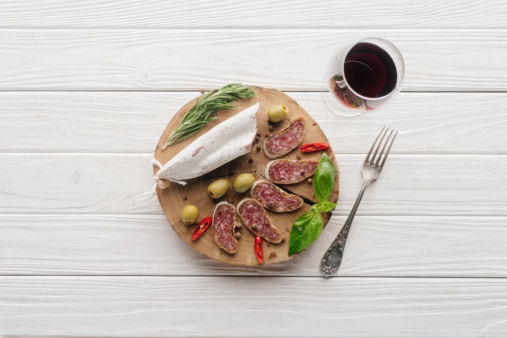 Blick von oben auf Gabel, Glas Rotwein und verschiedene Fleischsnacks auf weißem Holztisch - Foto, Bild