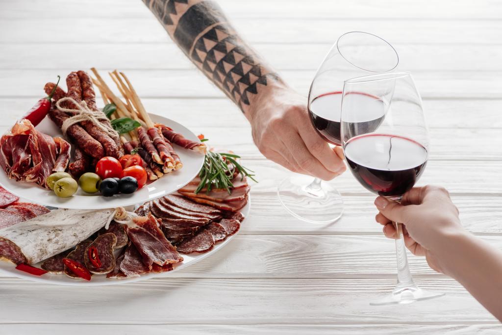 Teilansicht von Paar klimpernde Gläser mit Rotwein an weißer Tischplatte mit Fleischvorspeisen - Foto, Bild