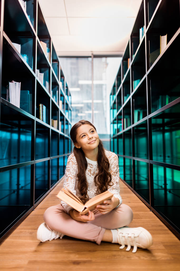 hermosa colegiala sonriente sosteniendo libro y mirando hacia otro lado mientras está sentado en el suelo en la biblioteca
 - Foto, Imagen