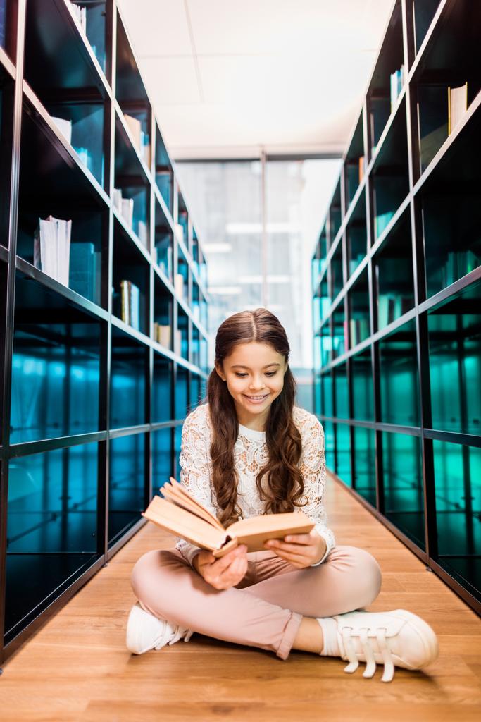 krásnou usměvavou školačka sedí na podlaze a čtení knih v knihovně - Fotografie, Obrázek