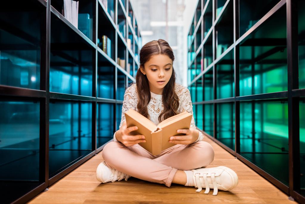 hermosa colegiala enfocada sentada en el suelo y leyendo libro en la biblioteca
   - Foto, imagen