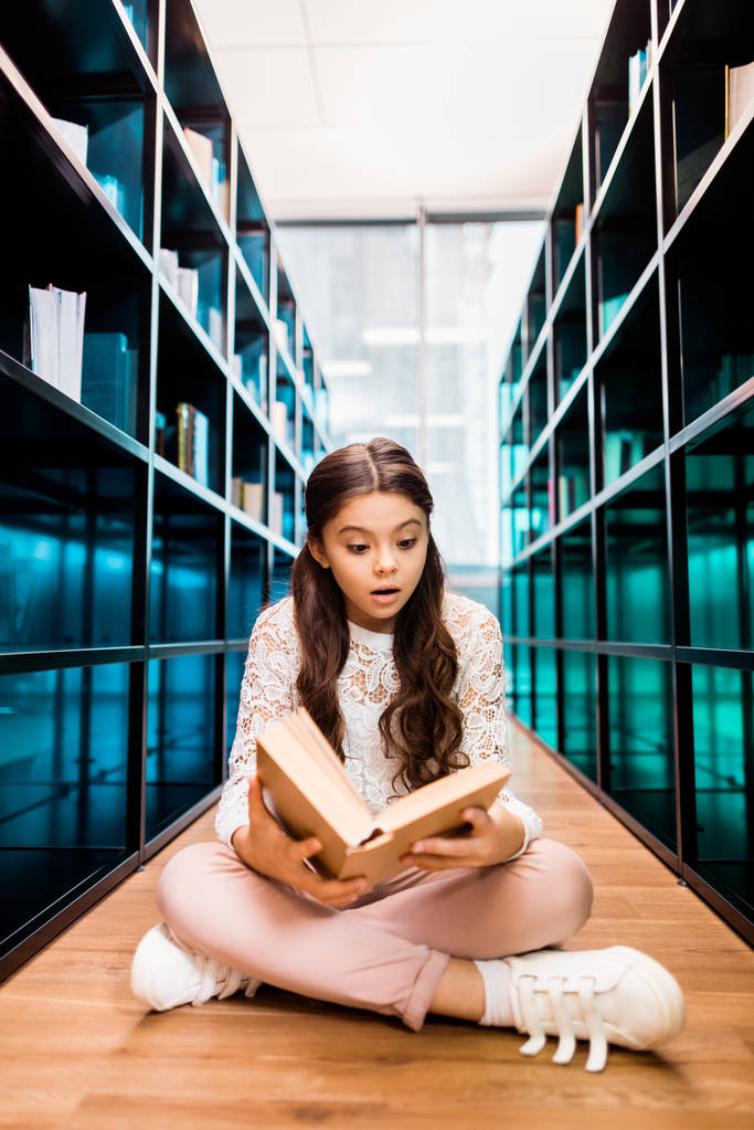 Urocza uczennica szoku, siedząc na podłodze i czytanie książki w bibliotece  - Zdjęcie, obraz