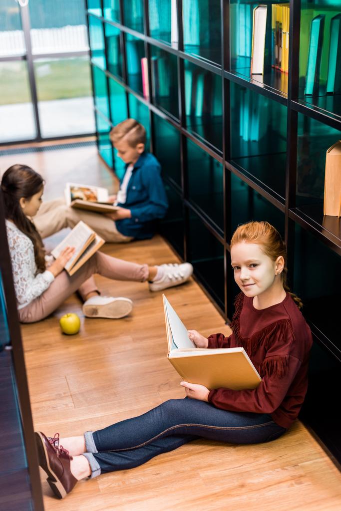 высокий угол обзора очаровательного школьника, держащего книгу и улыбающегося перед камерой во время чтения с одноклассниками в библиотеке
 - Фото, изображение