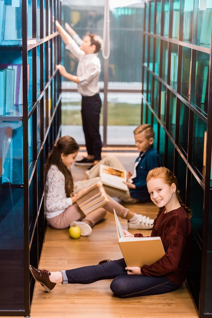 vista de ángulo alto de adorables escolares leyendo libros en el suelo en la biblioteca
  - Foto, Imagen