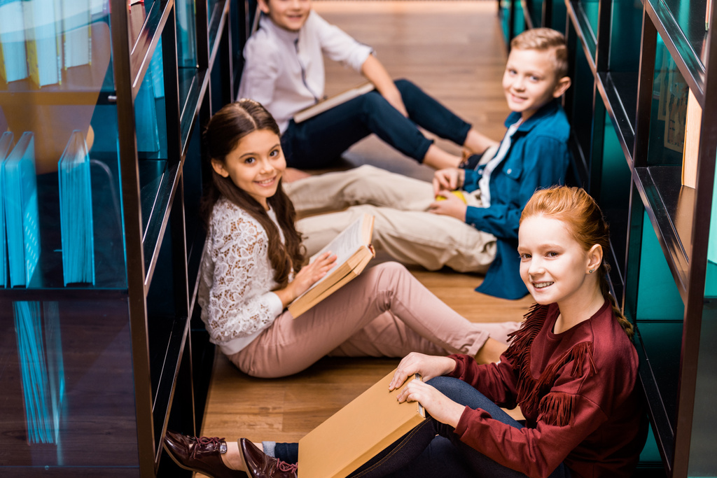 hoge hoekmening van klasgenoten met boeken zittend op de vloer en glimlachend in de camera in de bibliotheek   - Foto, afbeelding