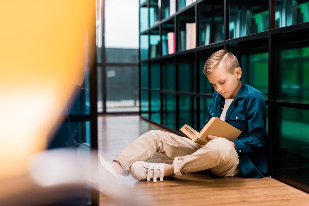 lindo centrado niño leyendo libro y sentado en el suelo en la biblioteca
   - Foto, imagen