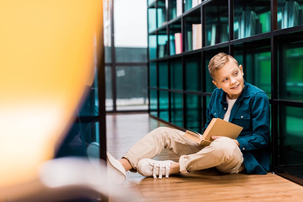 schattige lachende jongen boek lezen en bekijken weg zittend op de vloer in bibliotheek   - Foto, afbeelding