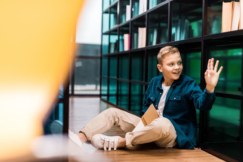 cute smiling boy holding book and waving hand while sitting on floor in library  - Photo, Image