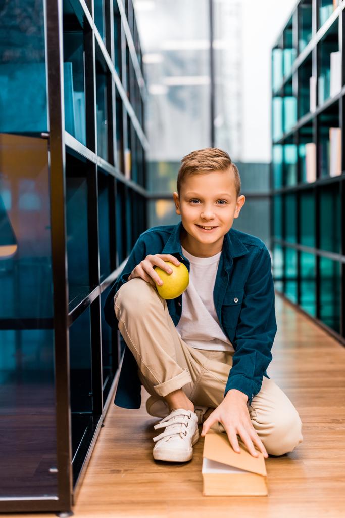 очаровательный школьник держит яблоко и улыбается в камеру, сидя на полу в библиотеке
  - Фото, изображение