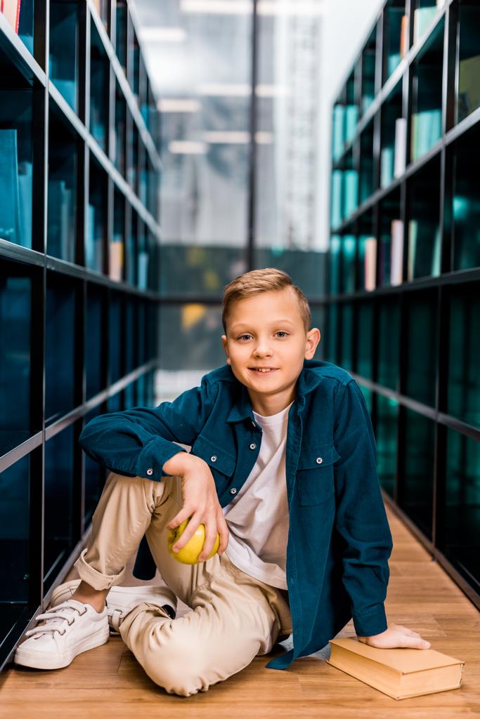 roztomilý kluk drží jablko a usmívá se na kameru, zatímco sedí na podlaze v knihovně  - Fotografie, Obrázek