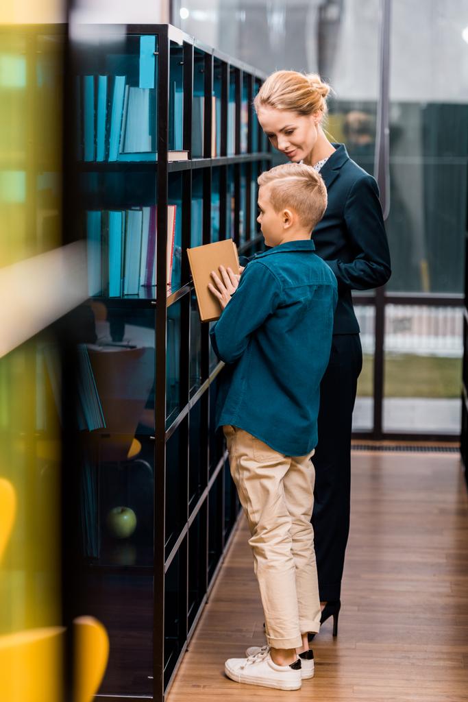 sonriente joven bibliotecaria mirando niño sosteniendo libro en la biblioteca
  - Foto, Imagen