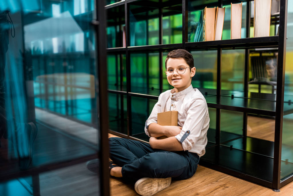 cute happy schoolboy in eyeglasses holding book and sitting on floor in library  - Photo, Image