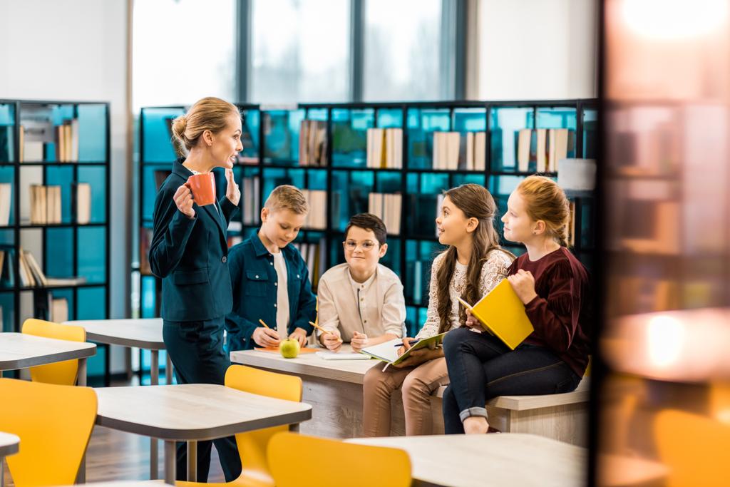 young female librarian and schoolkids talking together in library - Photo, Image