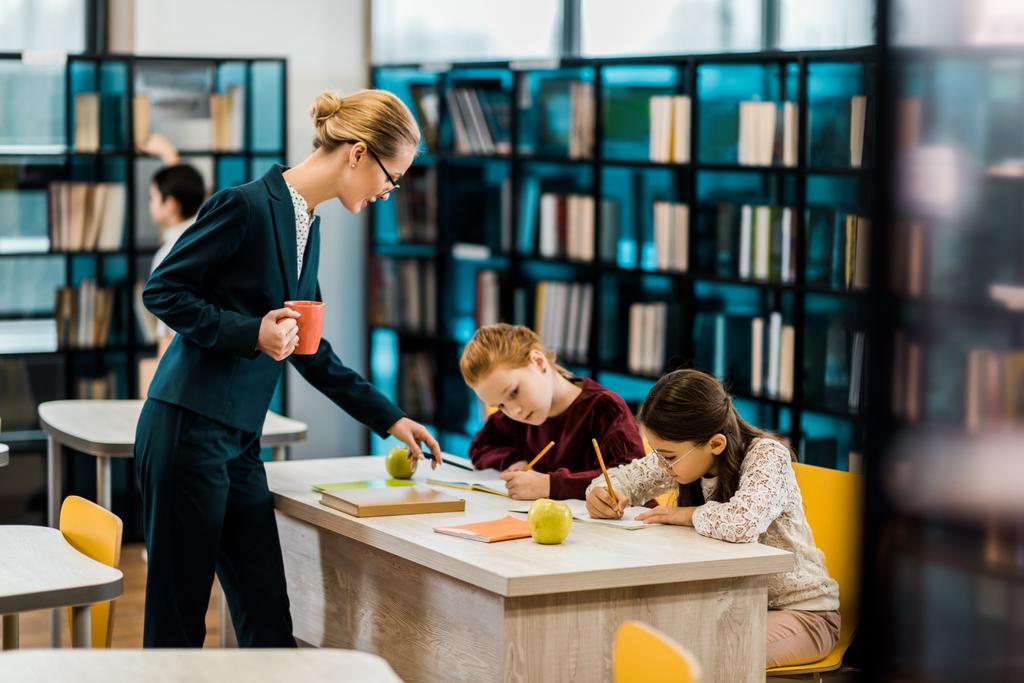 young female teacher holding cup and looking at schoolchildren studying in library    - Photo, Image