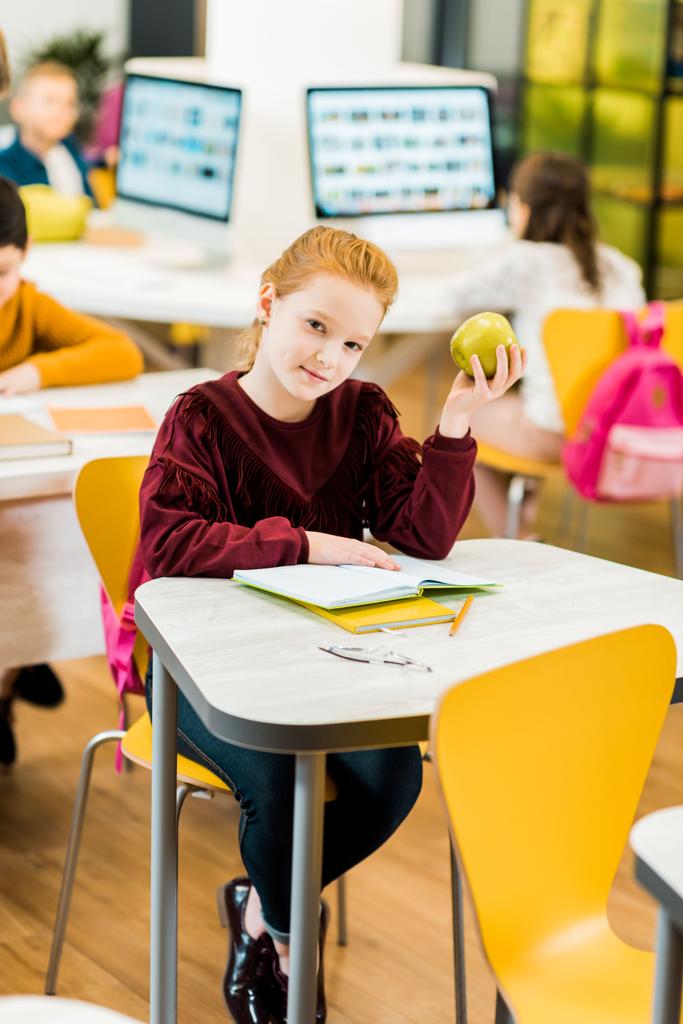 entzückendes Schulkind, das einen Apfel in der Hand hält und in die Kamera schaut, während es mit Mitschülern in der Bibliothek sitzt - Foto, Bild