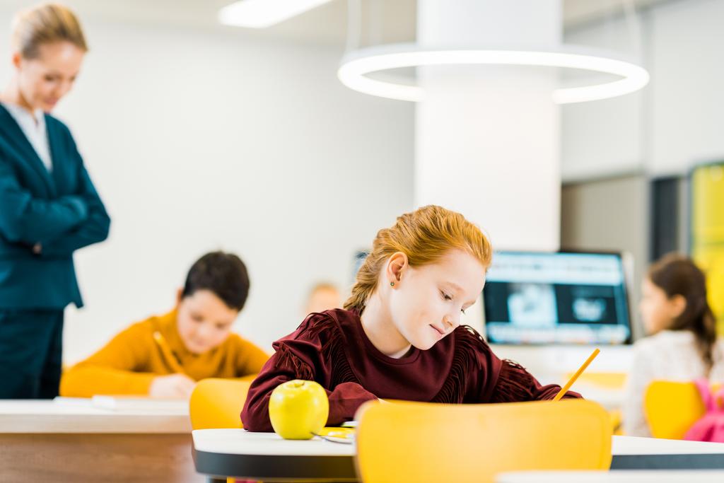 Lehrerin sieht entzückende Schulkinder, die am Schreibtisch schreiben und lernen - Foto, Bild