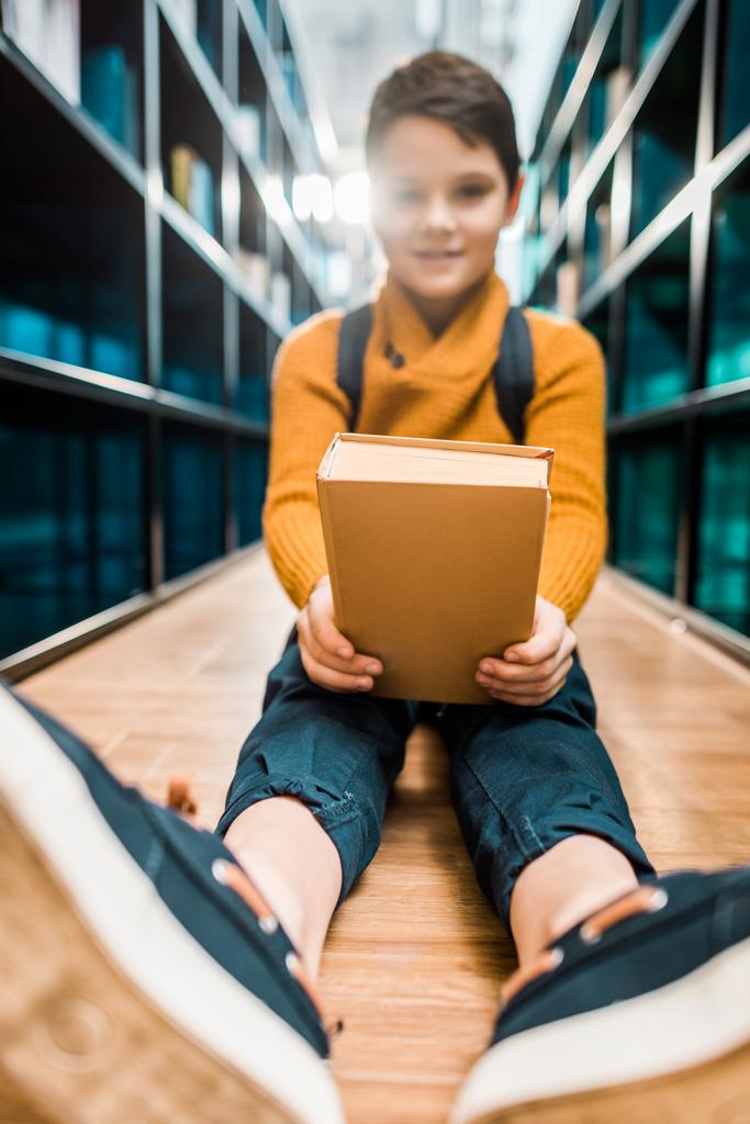 schoolboy holding book and smiling at camera while sitting on floor in library - Photo, Image