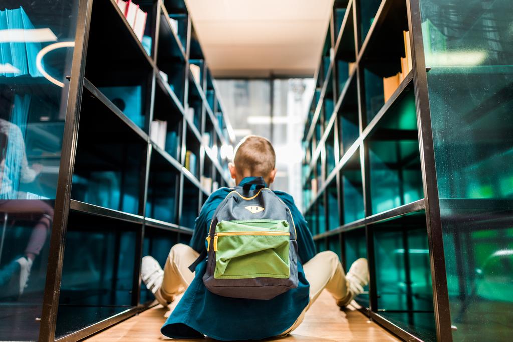 πίσω όψη του αγοριού με σακίδιο που κάθεται στο πάτωμα στη βιβλιοθήκη  - Φωτογραφία, εικόνα