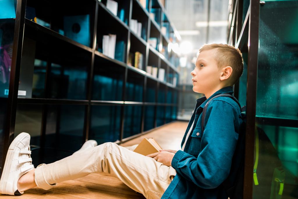 side view of cute schoolboy holding book and looking away while sitting on floor in library  - Photo, Image