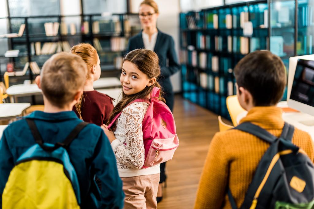 чарівна школярка з рюкзаком посміхається на камеру під час відвідування бібліотеки з однокласниками
 - Фото, зображення