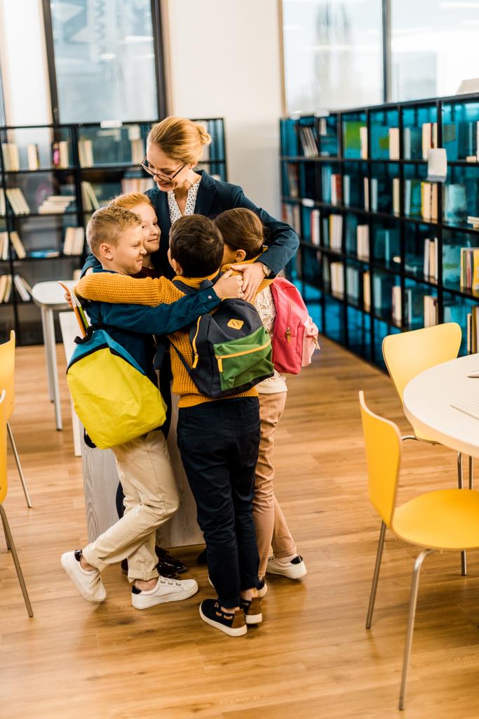 crianças felizes com mochilas abraçando bibliotecário na biblioteca
  - Foto, Imagem