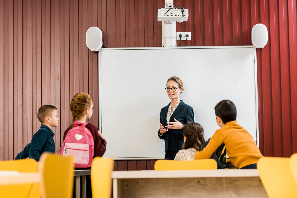jonge leraar permanent in de buurt van interactieve whiteboard en schoolkinderen kijken met rugzakken  - Foto, afbeelding