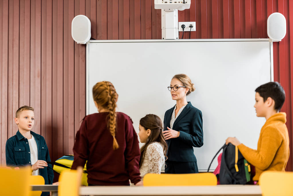 νεαρή δασκάλα, στέκεται κοντά το διαδραστικό πίνακα και κοιτάζοντας τους μαθητές με Σακίδια  - Φωτογραφία, εικόνα