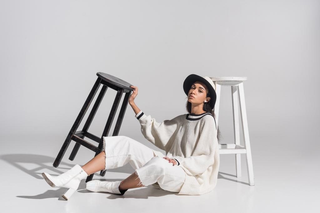 attractive african american girl in stylish white clothes and hat sitting near black and white chairs on white - Photo, Image