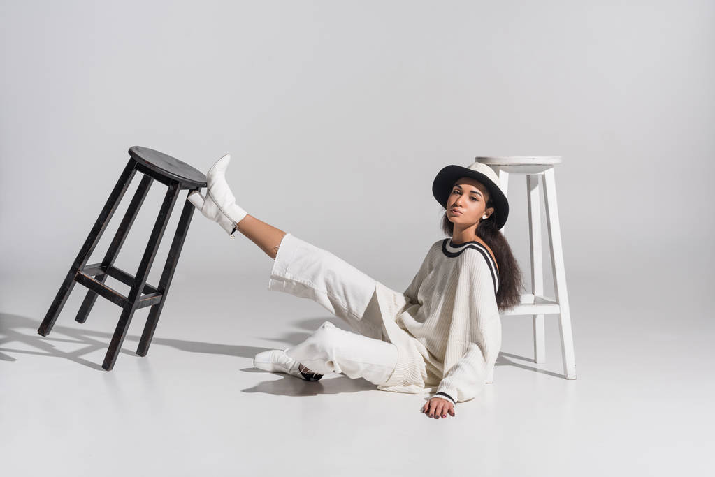 attractive african american young woman in stylish white clothes and hat sitting near black and white chairs on white - Photo, Image