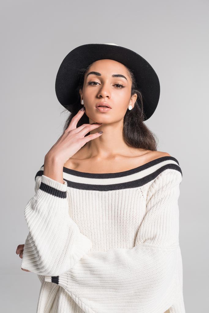attractive african american girl in stylish white sweater and hat touching face and looking at camera isolated on white - Photo, Image
