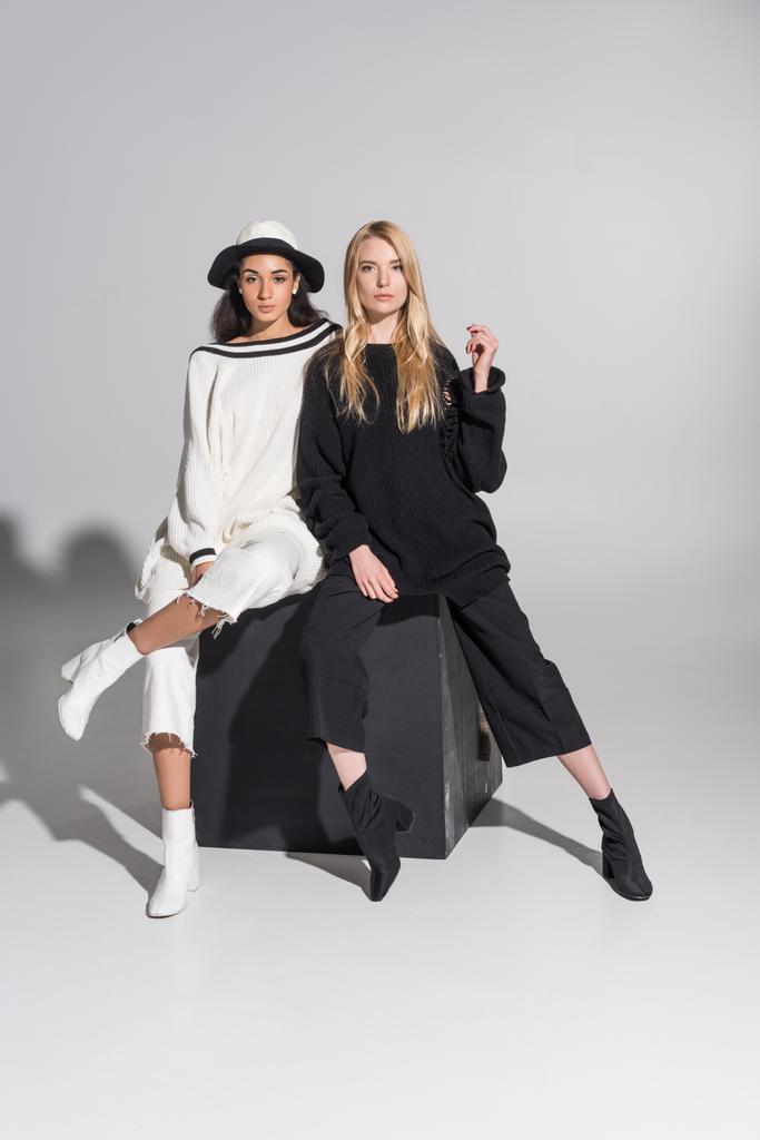 wunderschöne multikulturelle Models in modischen schwarz-weißen Kleidern sitzen auf einem schwarzen Würfel und schauen in die Kamera auf weiß - Foto, Bild