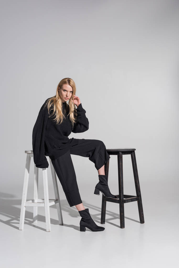 bela mulher loira caucasiana em roupas pretas sentado em cadeiras e olhando para a câmera no branco
 - Foto, Imagem