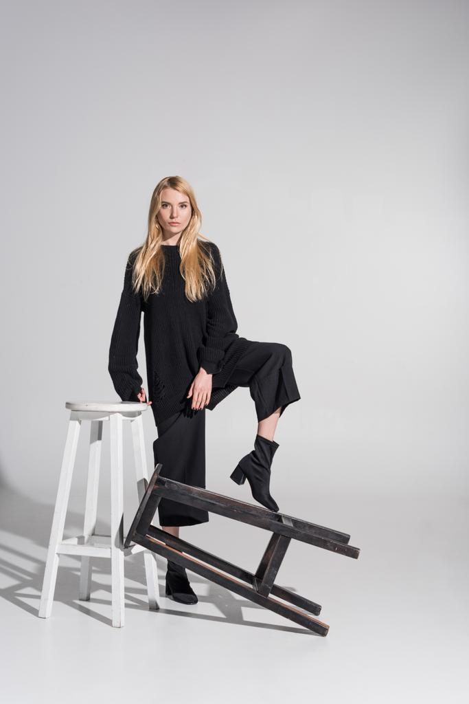 atractiva mujer rubia caucásica en ropa negra de pie y poner la pierna en la silla en blanco
 - Foto, imagen