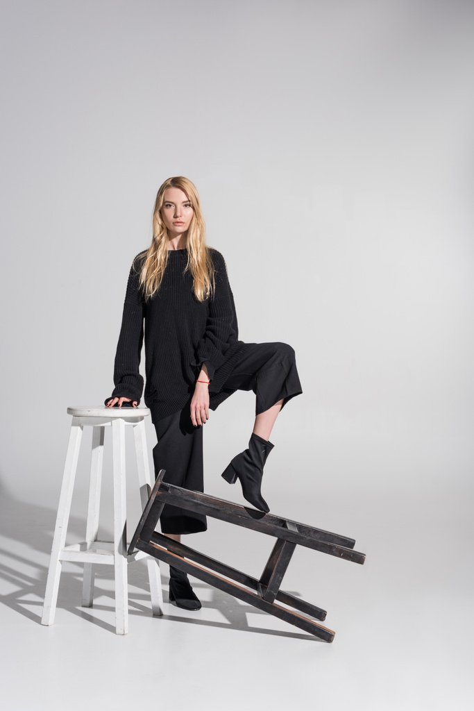 bella donna bionda caucasica in abiti neri in piedi e mettendo la gamba sulla sedia su bianco
 - Foto, immagini