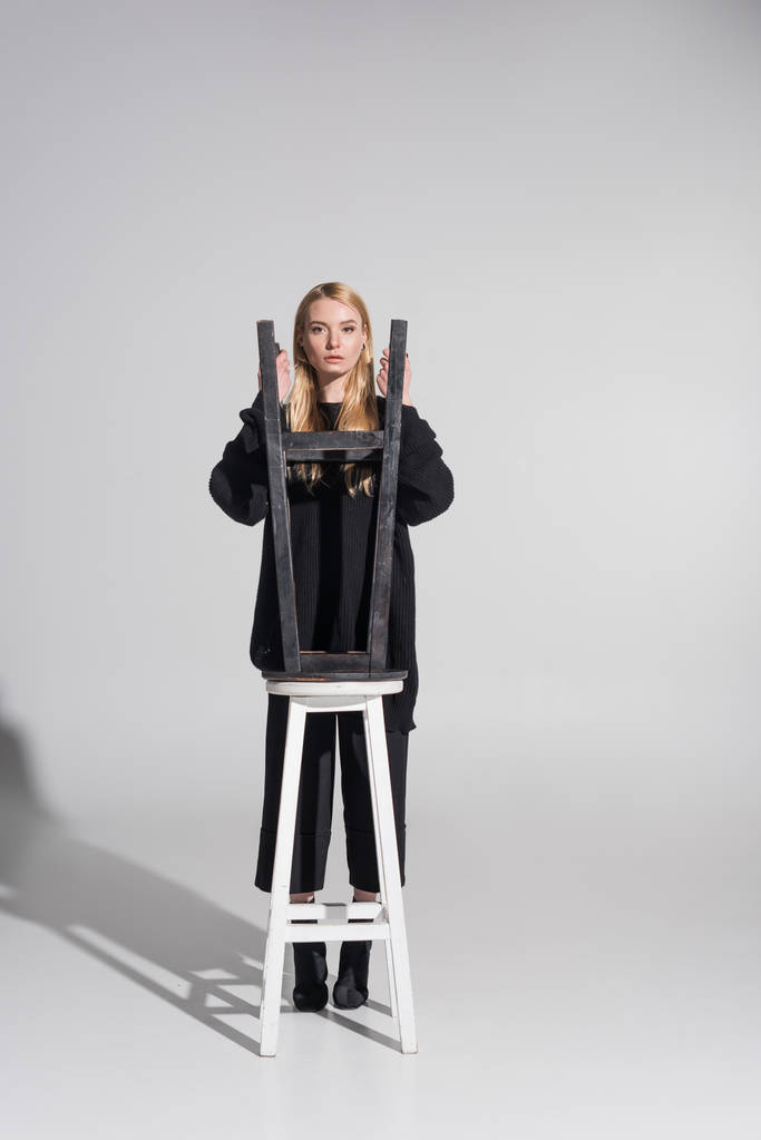 bela mulher loira caucasiana em roupas pretas olhando para a câmera através de cadeiras em branco
 - Foto, Imagem