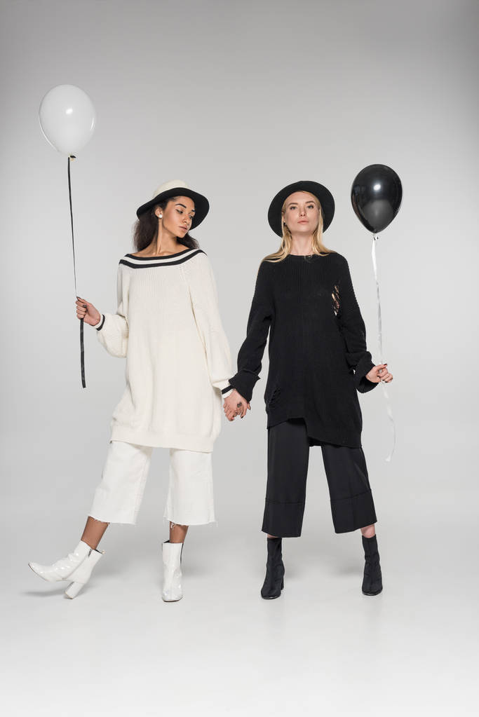 hermosa pareja de lesbianas multiétnicas en ropa blanca y negra cogidas de la mano y dos globos en blanco
 - Foto, Imagen