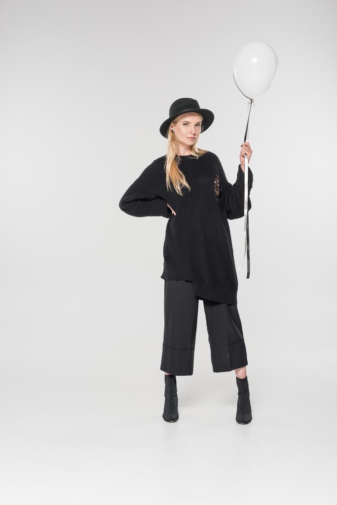 attractive caucasian blonde woman in stylish black clothes and hat holding balloons on white and looking at camera - Photo, Image