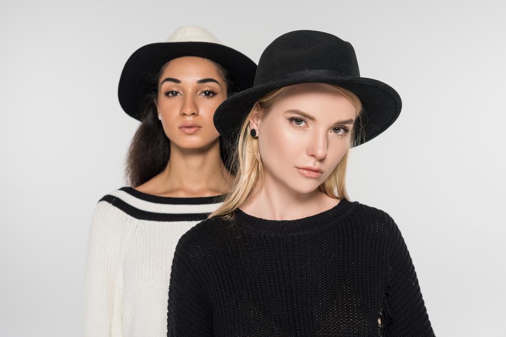 портрет красивых мультикультурных женщин в черно-белой одежде и шляпах, смотрящих в камеру, изолированную на белом
 - Фото, изображение