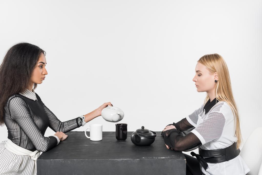 вид сбоку привлекательных мультиэтнических женщин в черно-белой одежде, сидящих за столом с чайниками и чашками изолированы на белом
 - Фото, изображение