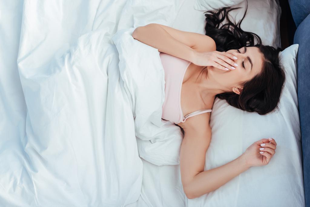 высокий угол зрения на молодую женщину зевая и закрывая рот рукой на кровати в утреннее время дома
 - Фото, изображение