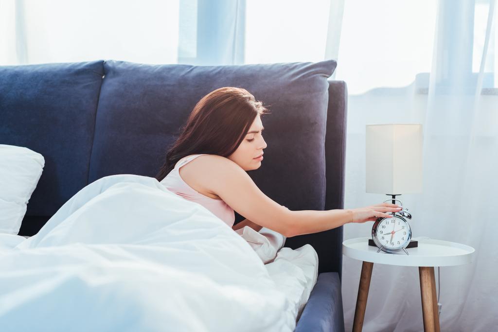 вид збоку молодої жінки, що вимикає будильник у спальні в ранковий час вдома
 - Фото, зображення