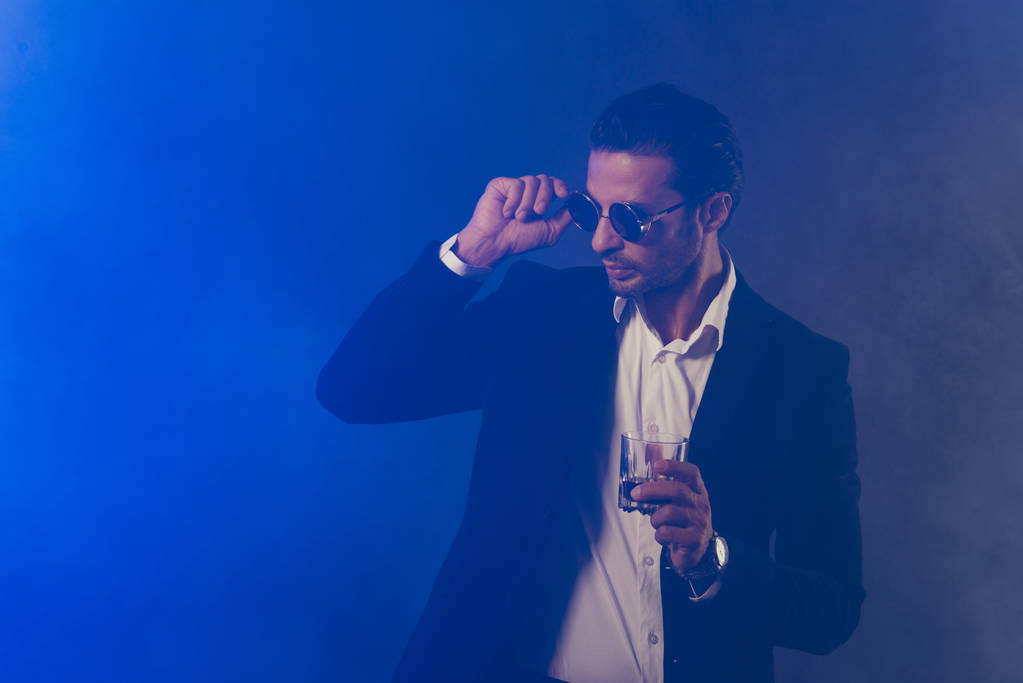 Портрет хороший прохладный привлекательный красивый уверенный в себе мужчина в черной куртке трогательные очки очки держа стекло весело изолированы на темно-синий дым фоне
 - Фото, изображение