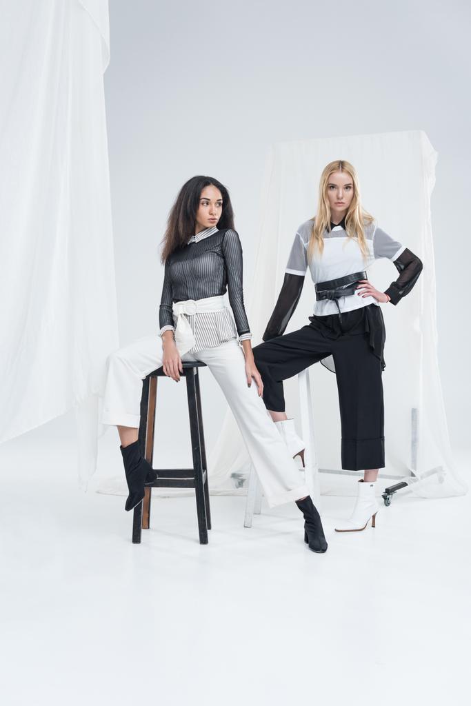attractive multiethnic women in fashionable black and white clothes posing on chairs on white - Photo, Image