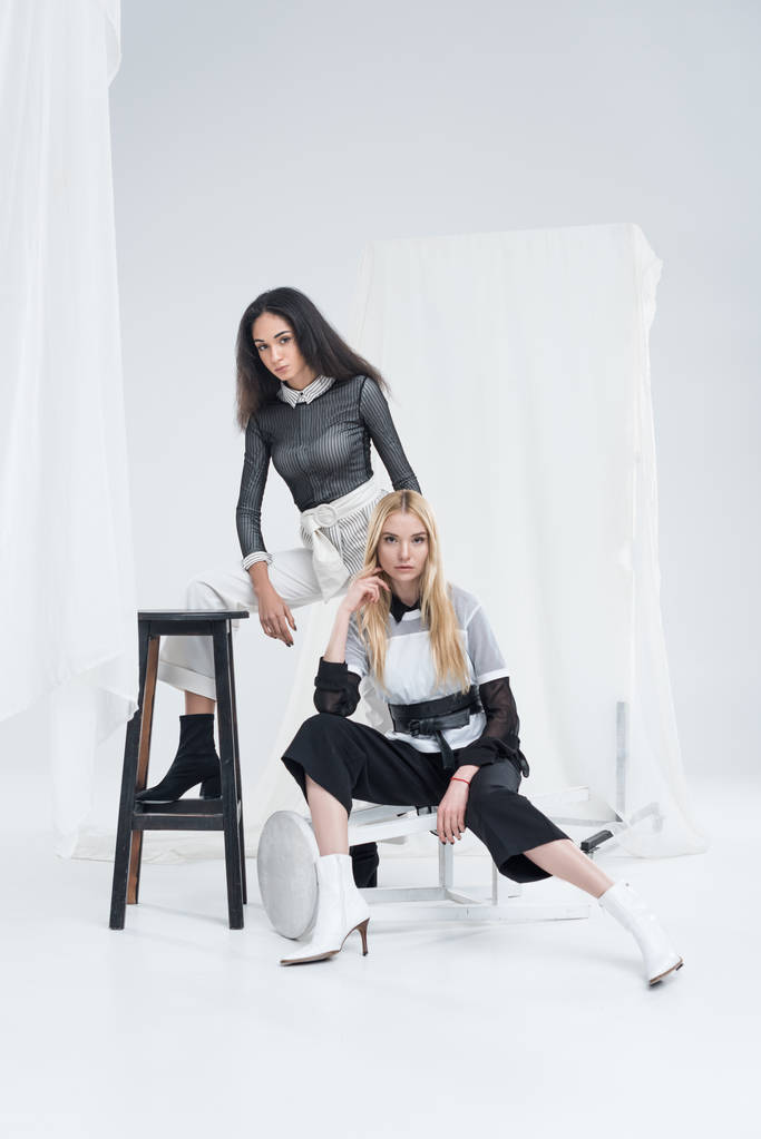 ελκυστικές γυναίκες πολυεθνική σε μοντέρνα ρούχα σε μαύρο και άσπρο που παρουσιάζουν στις καρέκλες κοντά σε τούλι σε λευκό - Φωτογραφία, εικόνα
