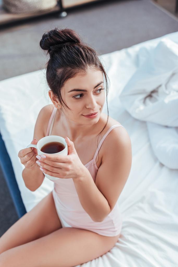 μεγάλη γωνία προβολής ευτυχισμένη νεαρή γυναίκα κάθεται με φλιτζάνι καφέ στο κρεβάτι κατά τη διάρκεια του χρόνου το πρωί στο σπίτι - Φωτογραφία, εικόνα