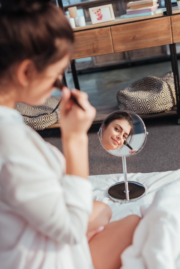 επιλεκτική εστίαση του όμορφο κορίτσι εφαρμογή μακιγιάζ με πινέλο και κοιτάζοντας καθρέφτη στο κρεβάτι στο σπίτι - Φωτογραφία, εικόνα