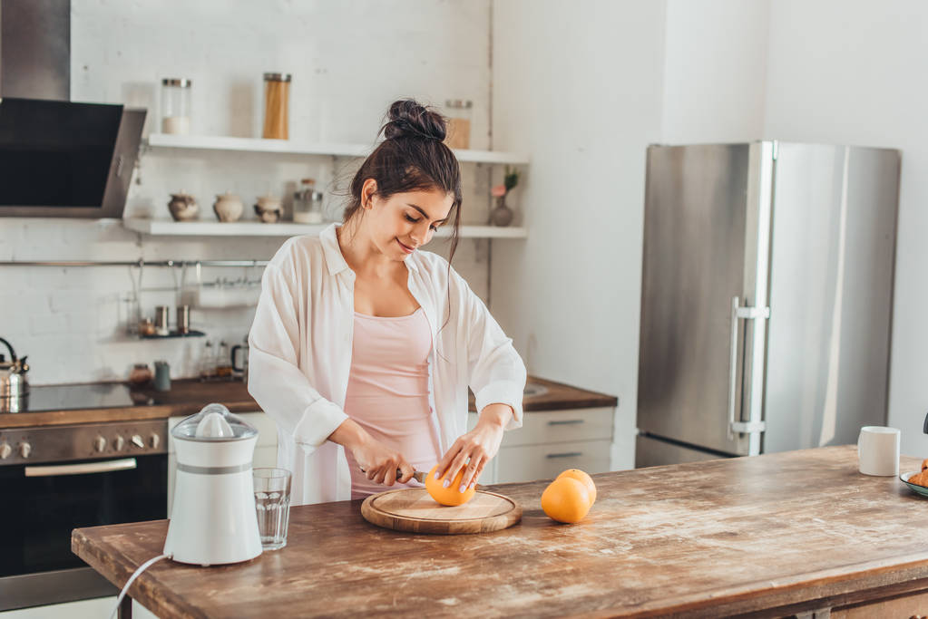 mulher sorrindo cortando laranja por faca na placa de madeira na cozinha
 - Foto, Imagem