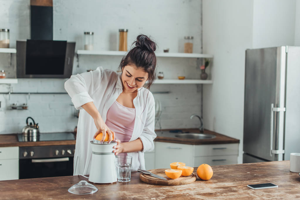 χαμογελαστό κορίτσι κάνει φρέσκο χυμό πορτοκάλι με αποχυμωτή στο ξύλινο τραπέζι στην κουζίνα - Φωτογραφία, εικόνα